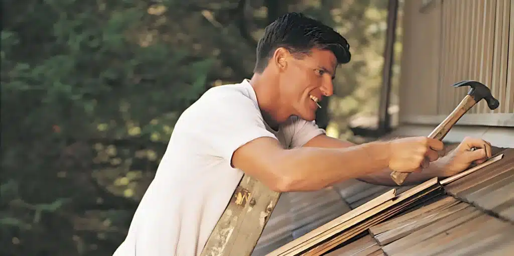 DIY vs. Professional Roof Repairs: Pros and Cons 1 A man in a white t-shirt is on a ladder, using a hammer to nail shingles on a roof, showcasing the spirit of DIY vs. professional roof repairs. Trees are visible in the background.