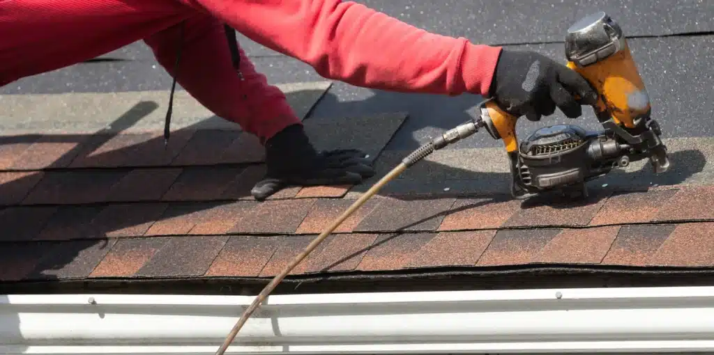 DIY vs. Professional Roof Repairs: Pros and Cons 3 A person in a red long-sleeve shirt and black gloves is using a nail gun to install asphalt shingles on a roof, showcasing the balance between DIY and professional roof repairs.