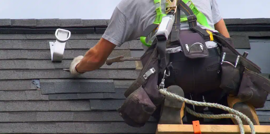 DIY vs. Professional Roof Repairs: Pros and Cons 2 A worker, embodying the essence of DIY vs. professional roof repairs, is wearing safety gear while hammering on a shingled roof. Tools secured on their belt and a rope for safety reflect their expertise and commitment to a job well done.