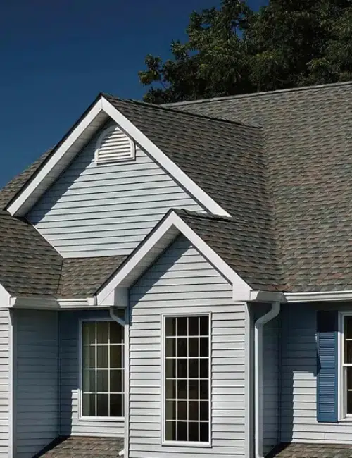 A house with light blue siding and dark shingles. It features multiple gables, a vent, and two windows. The sky is clear, and trees are partially visible in the background. A house with light blue siding and dark shingles. It features multiple gables, a vent, and two windows. The sky is clear, and trees are partially visible in the background.